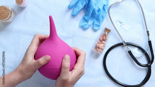 Doctor's hands holding a large pink enema on a table background with various objects, top view. Female doctor squeezing a pink rubber enema. Nurse or doctor's desk with a stethoscope, pills and gloves
