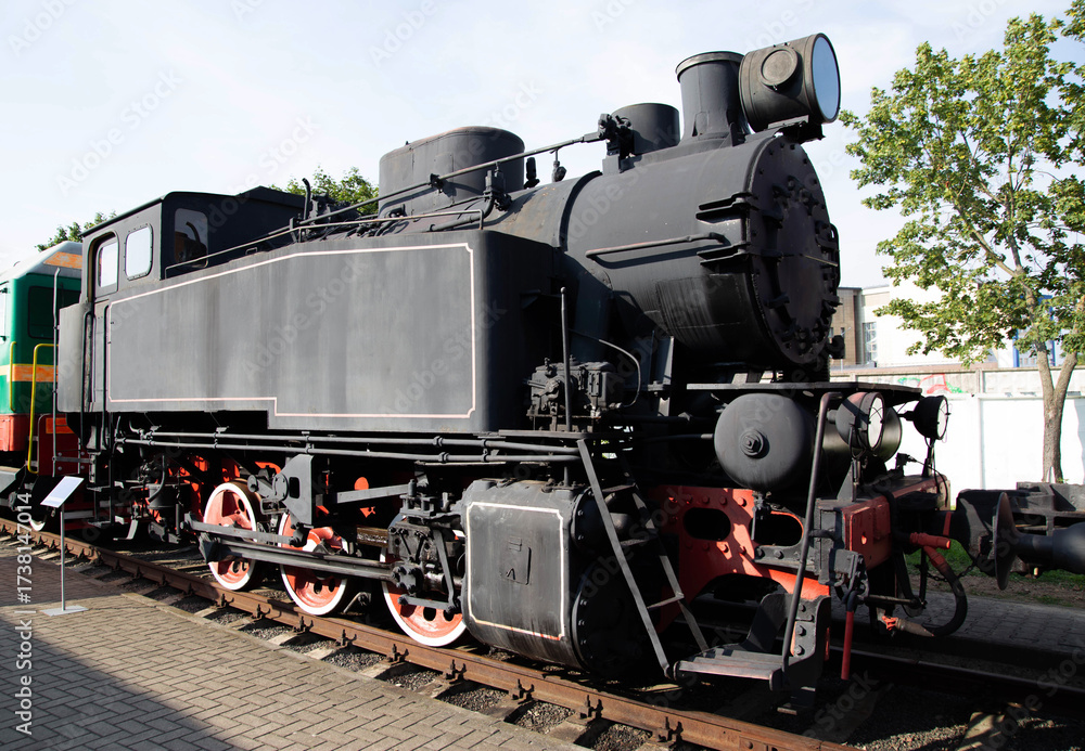 Naklejka premium Old steam locomotive on display at a railway museum during a sunny afternoon in a bustling city