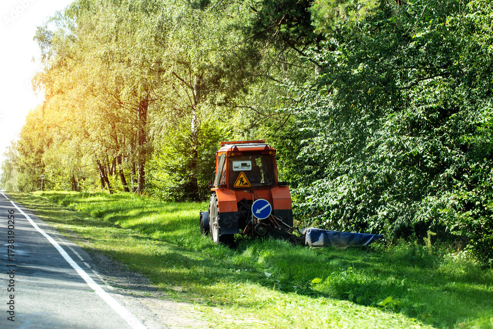 Obraz premium A special tractor with a mower for mowing grass on the side of the road against the background of a forest, industry. Copy space for text