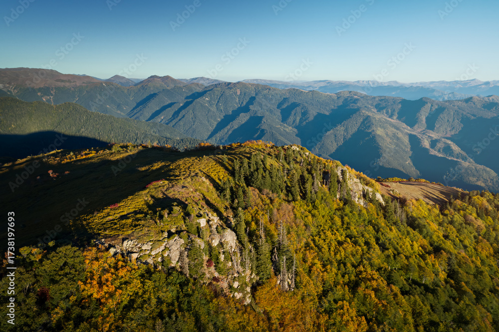 Fototapeta premium Golden sunlight highlights autumn foliage on a highland ridge, with distant mountain peaks fading into soft blue haze, Georgia.