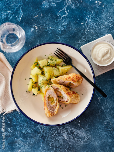 Tender chicken kiev is plated beside fluffy potatoes sprinkled with herbs, offering a tasty meal. A glass of water and a small bowl of sauce complete the setting