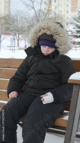 Lonely, sad boy with cast on arm swings on winter day. Vertical video of frustrated child who cant play with everyone due to injury.