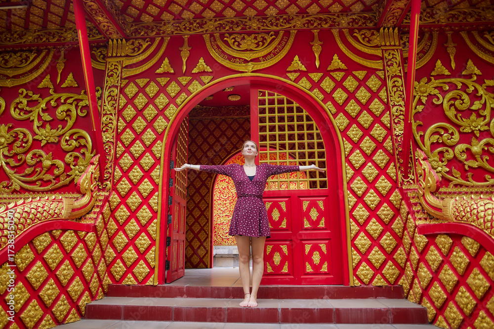 Obraz premium A Joyful Young Woman Poses Gracefully at the Entrance of a Vibrantly Decorated Temple with Rich Red and Golden Details