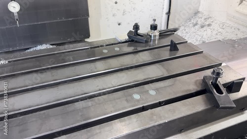 CNC mill T‑slot table with clamping kit: step block, strap clamp with stud and nut, T‑nut in the slot, and a combination wrench resting on the surface amid scattered chips and coolant marks