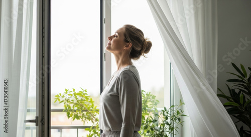 Free Download Serene woman stands near bright window in cozy home, enjoying fresh air and sunlight. space is adorned with green plants, creating peaceful and hygienic atmosphere for personal care - GFXLoad