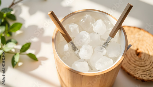 Ice bucket with ice cubes and tongs on wooden table with greenery  