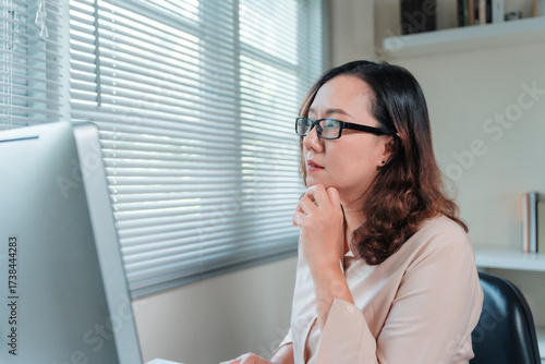Woman in glasses concentrating, working on desktop computer at office desk, reflecting and planning new business ideas