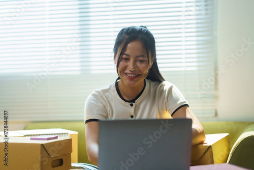 Young Asian woman smiling, working on laptop, preparing parcels for shipping in a home office, running a small business