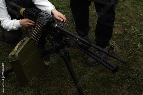 Person handling mounted machine gun with ammunition belt on grass, assisted by another. Military-style firearm setup in outdoor environment