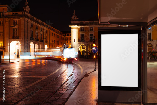 Blank vertical billboard mock-up at a bus stop in Lisbon at night, illuminated by warm city lights and blurred tram motion on wet cobblestone tracks, perfect for urban advertising design