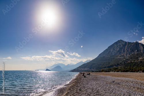 Fototapeta Naklejka Na Ścianę i Meble -  Sarısu Beach in winter backlight with people fishing and relaxing on pebble shore, forested mountains in haze under sunny Mediterranean sky. Antalya, Turkey.

