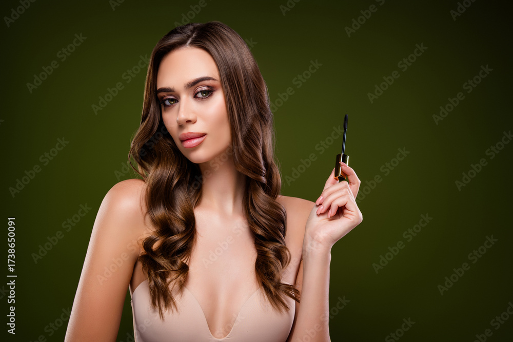 Obraz premium Elegant young woman with wavy hair holds mascara against a green background, showcasing beauty