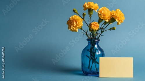 Yellow carnations in a blue vase with blank card on blue background for a greeting