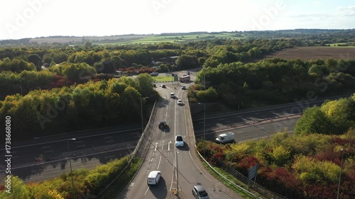 An aerial shot that focuses on an overpass bridge carrying road traffic over a motorway, leading into a distant roundabout, set within a landscape of abundant forest and fields.