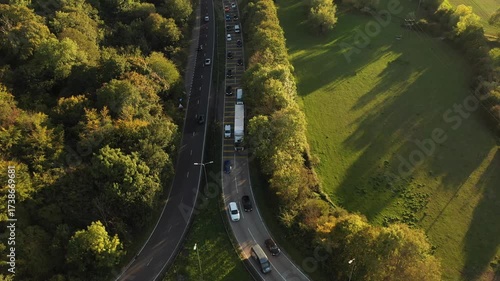 An aerial shot focusing on a road segment where a lane from a side road merges with the main dual carriageway, illustrating the flow of traffic with a long queue in one direction, framed by trees and 