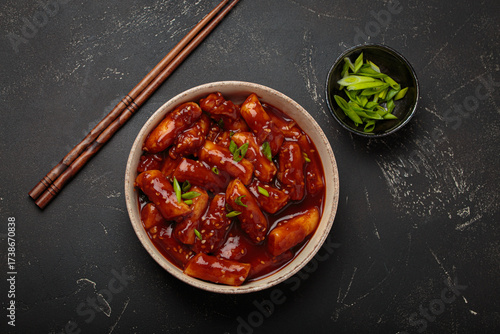 Stir fried rice cakes tteokbokki simmered in spicy sauce, garnished with green onions and sesame seeds, served on a black background with chopsticks top view. Popular dish of Korea