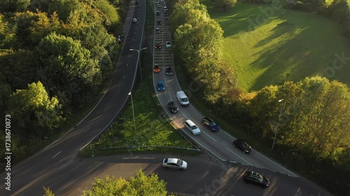 An aerial top-down view of a dual carriageway merge/exit point, with heavy traffic slowing and backed up on the main road, surrounded by vibrant green trees and fields under warm sunlight.
