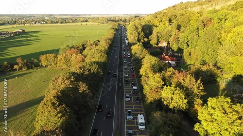 A steady, high-angle aerial view of a dual carriageway road with a pronounced traffic jam on one side, flanked by a wooded hillside and a wide, open field, with a small house nestled into the trees.
