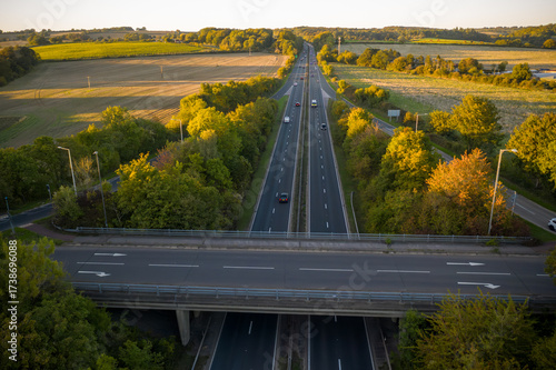A high-angle aerial view of a dual carriageway motorway passing underneath an overpass, flanked by lush green trees and open farmland bathed in the warm light of the setting sun.