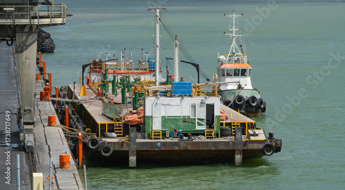 Fotografie Small tugboat is docked next to a barge with a structure and pipes, moored at a
