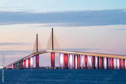 Sunshine Skyway Bridge over Tampa Bay in Florida, USA, at sunset. Red, White, and Blue lights shine under the bridge, and a pastel sky is visible behind the bridge. The water is dark navy below