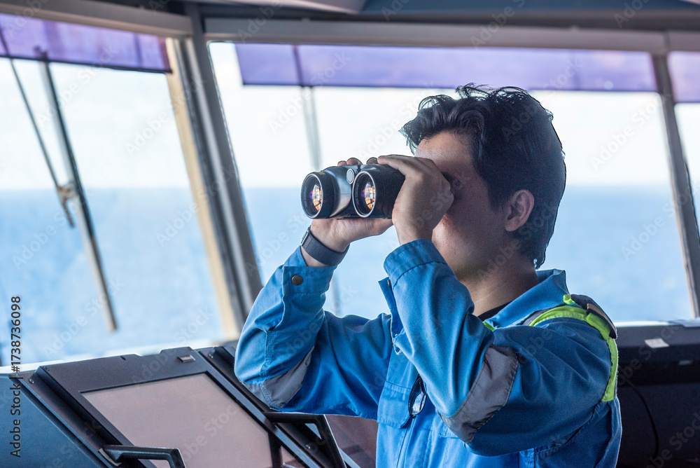 Obraz premium A male deck officer on the bridge of a merchant ship is looking through binoculars.