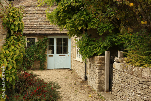 Cotswold stone cottage with blue door, gravel path leading to rustic entrance, overgrown walls, English village scene