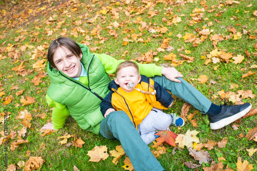 Fototapeta premium Teenage boy with his baby brother sitting on green grass with yellow maple leaves in autumn.