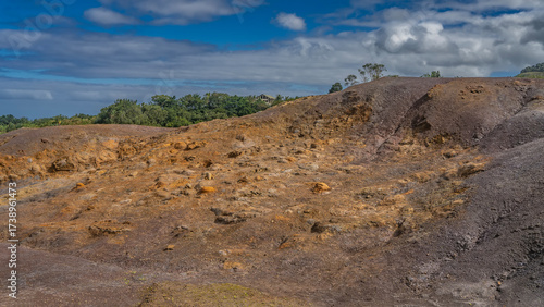 A unique geological landmark. 23 coloured earth of Mauritius. Multi-colored sand hill against a background of blue sky and clouds. Green vegetation in the distance. La Vallée Des Couleurs Nature Park