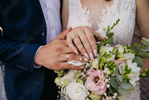 The bride and groom embrace each other on their wedding day and hold a bouquet of flowers on which they have placed their hands and are showing off their wedding rings