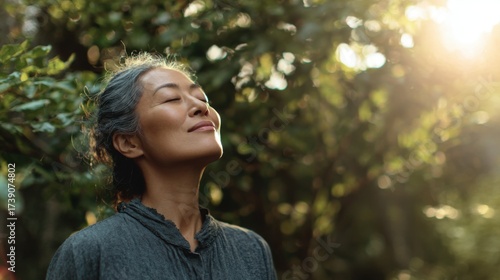 Medium view of a healthy Asian adult outdoors, standing in nature, closing eyes while taking a deep breath of fresh air, smiling with a calm and energized expression, posture open and relaxed
