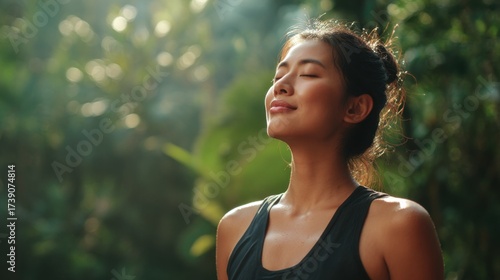 Medium view of a healthy Asian adult outdoors, standing in nature, closing eyes while taking a deep breath of fresh air, smiling with a calm and energized expression, posture open and relaxed

