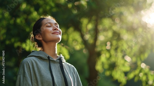 Medium view of a healthy Asian adult outdoors, standing in nature, closing eyes while taking a deep breath of fresh air, smiling with a calm and energized expression, posture open and relaxed
