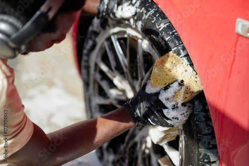 Obraz na plátně Black man wearing headphones cleaning car wheel with soapy sponge, focusing on s