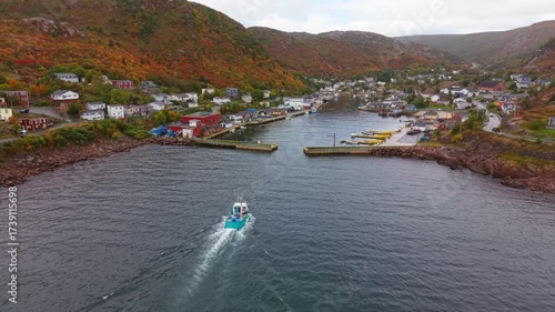 High drone shot over Petty Harbour, Newfoundland, in autumn captures a boat leaving the harbor, its wake trailing across calm waters, framed by rugged hills and colorful fall foliage.