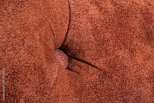 Close view of a brown fabric cushion with a central button detail. The textured surface and stitched folds highlight comfort and softness design typical in upholstered furniture. Selective focus.