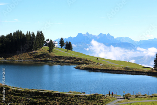 Scenic view of hikers walking along a lakeside path with mountains in the background during a sunny day in the Swiss Alps