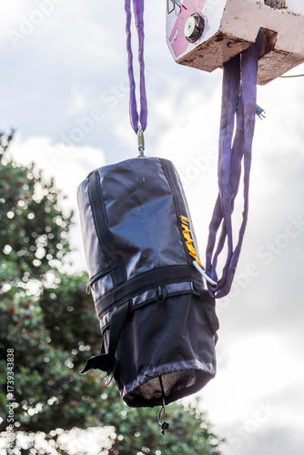 A black industrial bag secured with straps and suspended by a crane hook