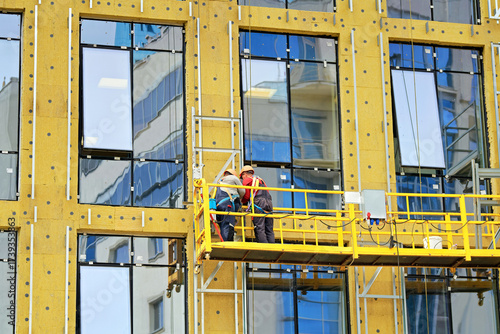 Building construction with workers on suspended scaffold hoist insulating exterior facade with glass panels and thermal insulation materials. Workers in safety gear work on suspended scaffold platform