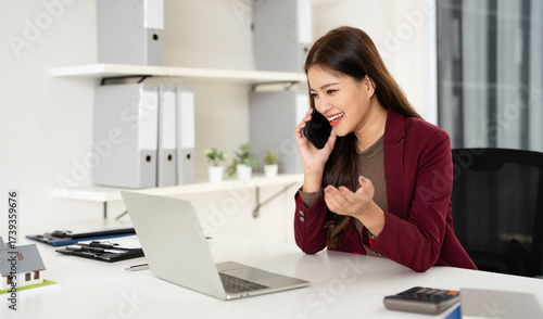 Confident asian business lady, broker real estate agent or manager, sitting at table in a modern office, working with laptop, talking to client or employees on smartphone, smiling.