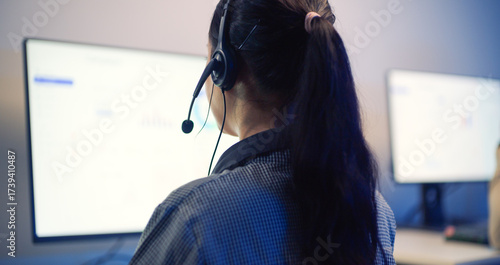 Close-up rear view of Asian woman wearing headset while working in call center office at night.