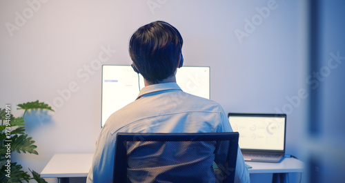 Back view asian male call center customer support with headset working late at night in modern office, using dual computer monitors for customer service, support and communication.