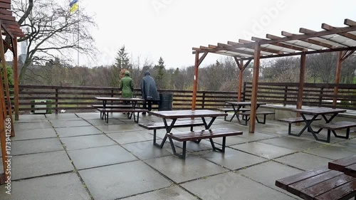 People on a wet outdoor terrace with picnic tables under an overcast sky, looking at a distant landscape with trees and the Ukrainian flag. A quiet, contemplative scene on a dreary day.