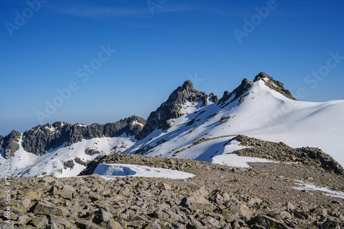 Snowy peaks and rocky terrain of Sierra de Gredos, Spain, beneath a vivid blue sky. The dramatic mountain landscape highlights rugged summits and pristine alpine environment.