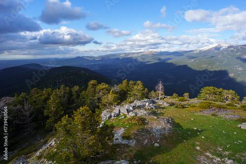 Rocky outcrop and pine forest at Cerro de la Camorca summit, Sierra de Guadarrama, Spain, with panoramic mountain views under vibrant blue sky and scattered clouds