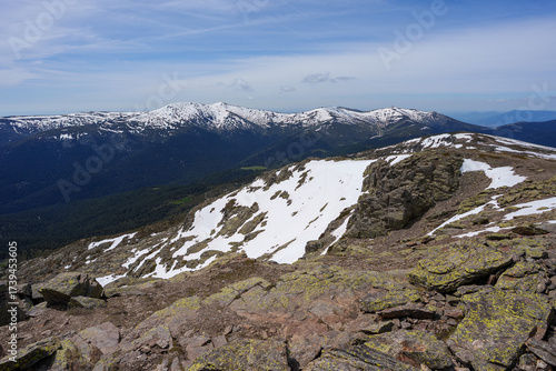 Stunning panoramic view from the Peñalara ridge in Sierra de Guadarrama. Rocky foreground with large patches of melting snow overlooking distant forested mountains under a blue sky.