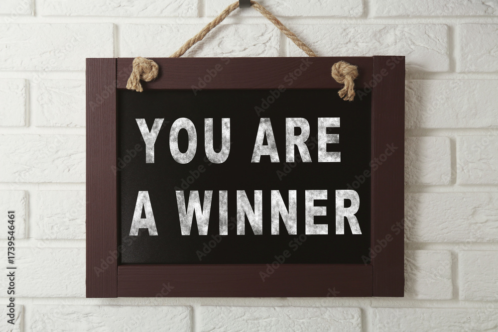 Fototapeta premium Chalkboard with phrase You Are A Winner hanging on white brick wall