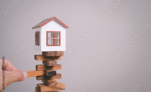 Canvas Print Close-up of hand removing a wooden block from a stack with small model house on top, uncertainty, decision-making, symbolizing risk in real estate investment