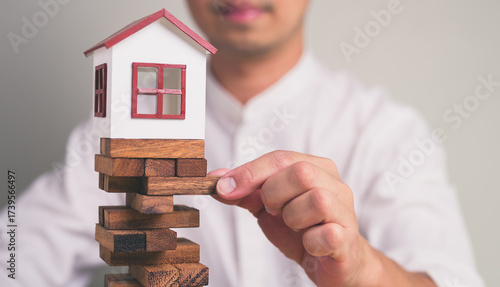 Businessman's hand removing a wooden block from a stack with small model house on top, uncertainty, decision-making, symbolizing risk in real estate investment
