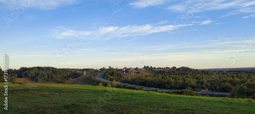 Picturesque view of rural landscape with winding road, green fields, and cluster of houses nestled among trees. Scene bathed in soft light, serene and peaceful atmosphere. Travel, nature and lifestyle